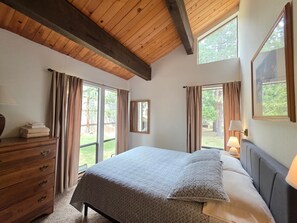 Ein Schlafzimmer mit Bett, Holzkommode, großen Fenstern und Blick ins Freie.