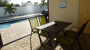 Un patio con una mesa y sillas, un cartel que indica una piscina, y vistas de la piscina y palmeras.