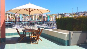 A terrace with a table and chairs under a large umbrella, overlooking a marina with colorful buildings in the background.