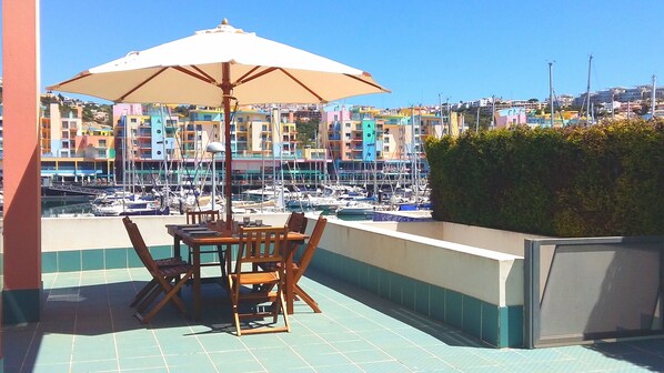 A terrace with a table and chairs under a large umbrella, overlooking a marina with colorful buildings in the background.