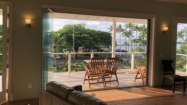Un vaste espace de vie avec une grande porte-fenêtre coulissante donnant sur un balcon aménagé avec du mobilier en bois et offrant une vue sur des palmiers et une étendue d’eau.