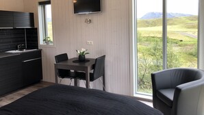 Una habitación moderna con una cama, una mesa de comedor con sillas, un televisor montado en la pared y un ventanal con vistas a un paisaje verde.