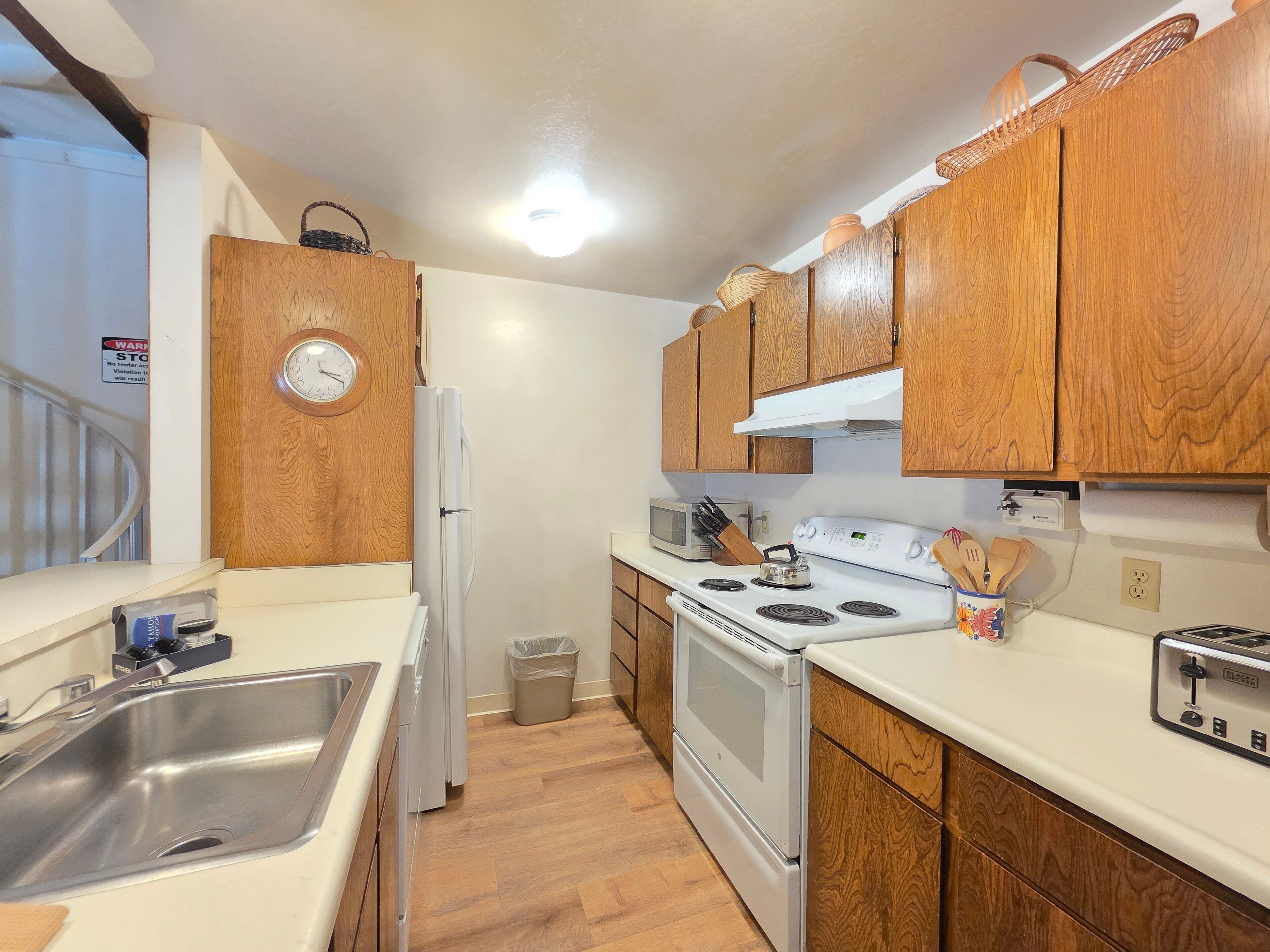 Una cocina compacta con armarios de madera, un lavamanos de acero inoxidable, un microondas, una estufa y un tostador.