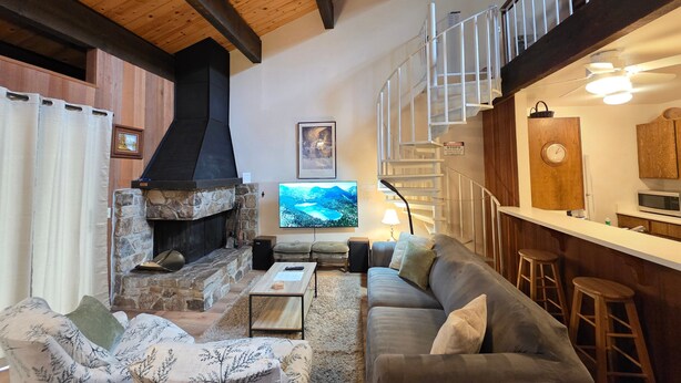 A living room with a stone fireplace, a sofa, a television, and a staircase leading to an upper level. - Kingwood Condo | Community Pool & Tennis Courts (Kings Beach)
