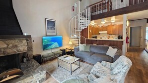A spacious living area with a stone fireplace, a large flat-screen TV, a gray sofa, and a staircase leading to an upper level. - Kingwood Condo | Community Pool & Tennis Courts (Kings Beach)