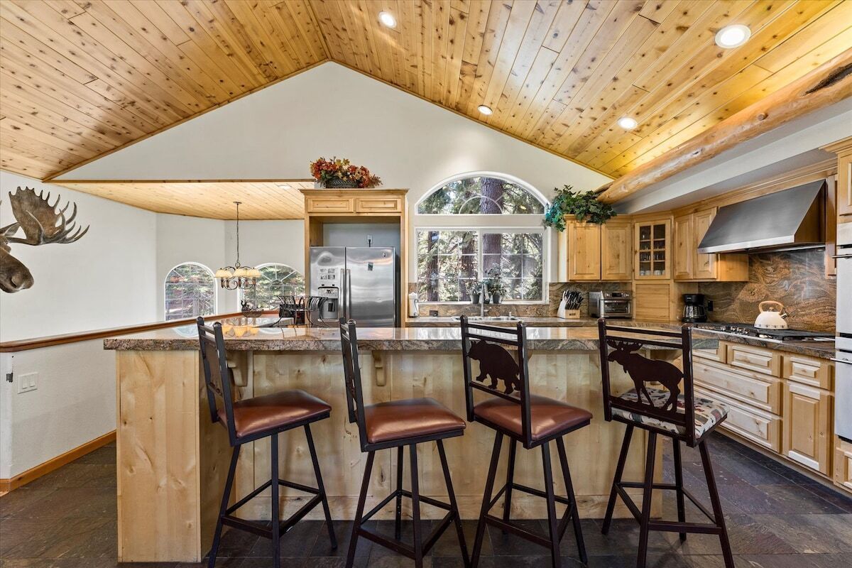 Una cocina amplia con armarios de madera, una isla grande con taburetes de bar, un refrigerador de acero inoxidable y vistas al exterior a través de un ventanal.