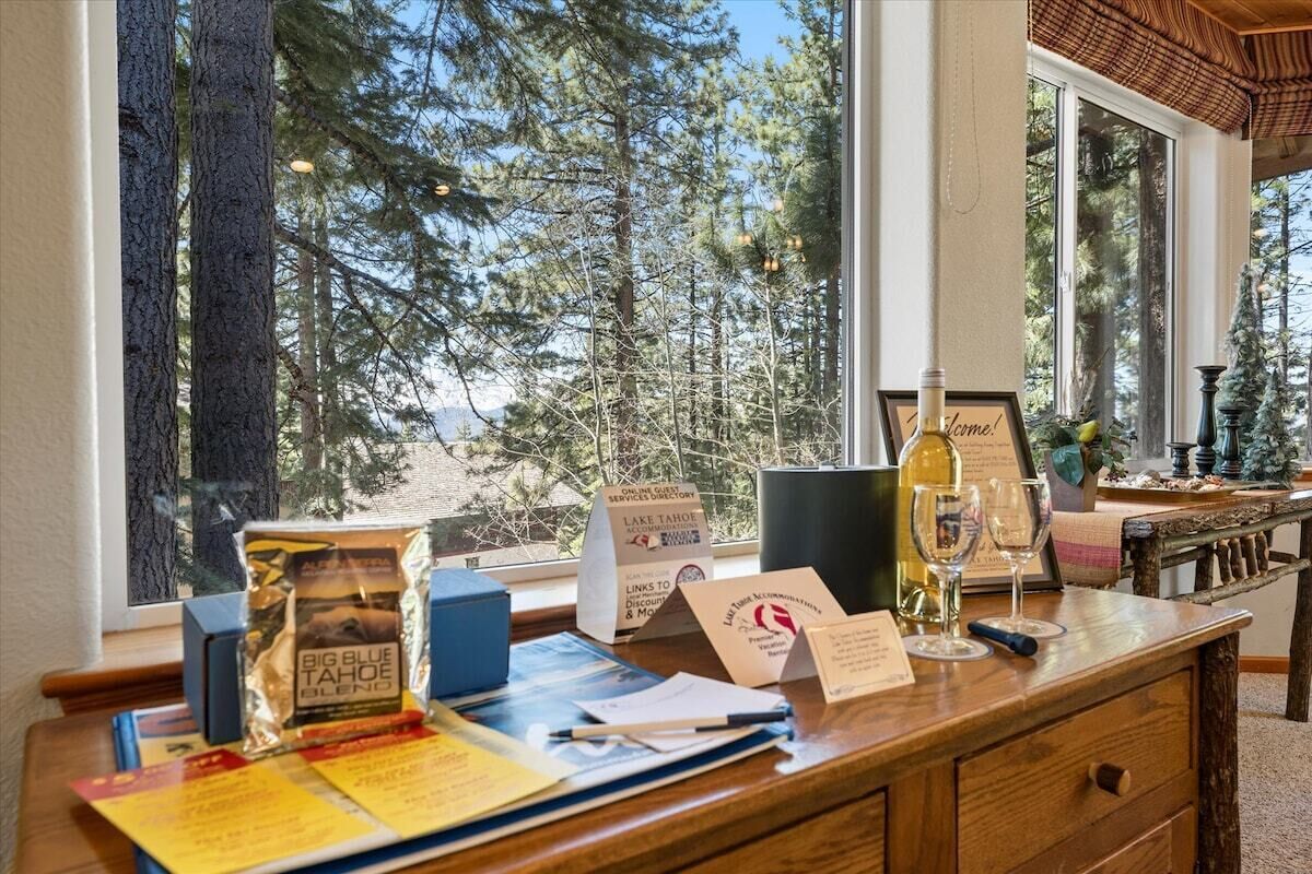 A wooden desk with documents, a wine bottle, and glasses, overlooking a forested area through large windows.
