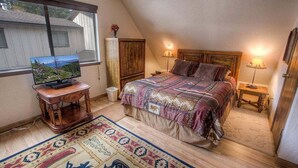 Ein Schlafzimmer mit einem großen Bett, zwei Nachttischlampen, einem Fernseher auf einem Holzständer und einem Fenster mit Blick auf ein Haus.