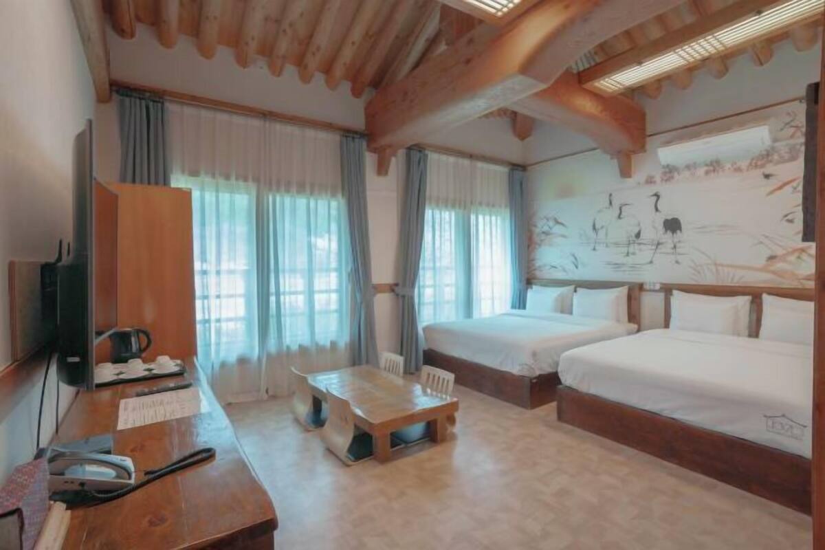 Ein Hotelzimmer mit einem Holztisch, zwei Betten, einem großen Fenster mit Vorhängen und einer Wand mit Kunstwerken.