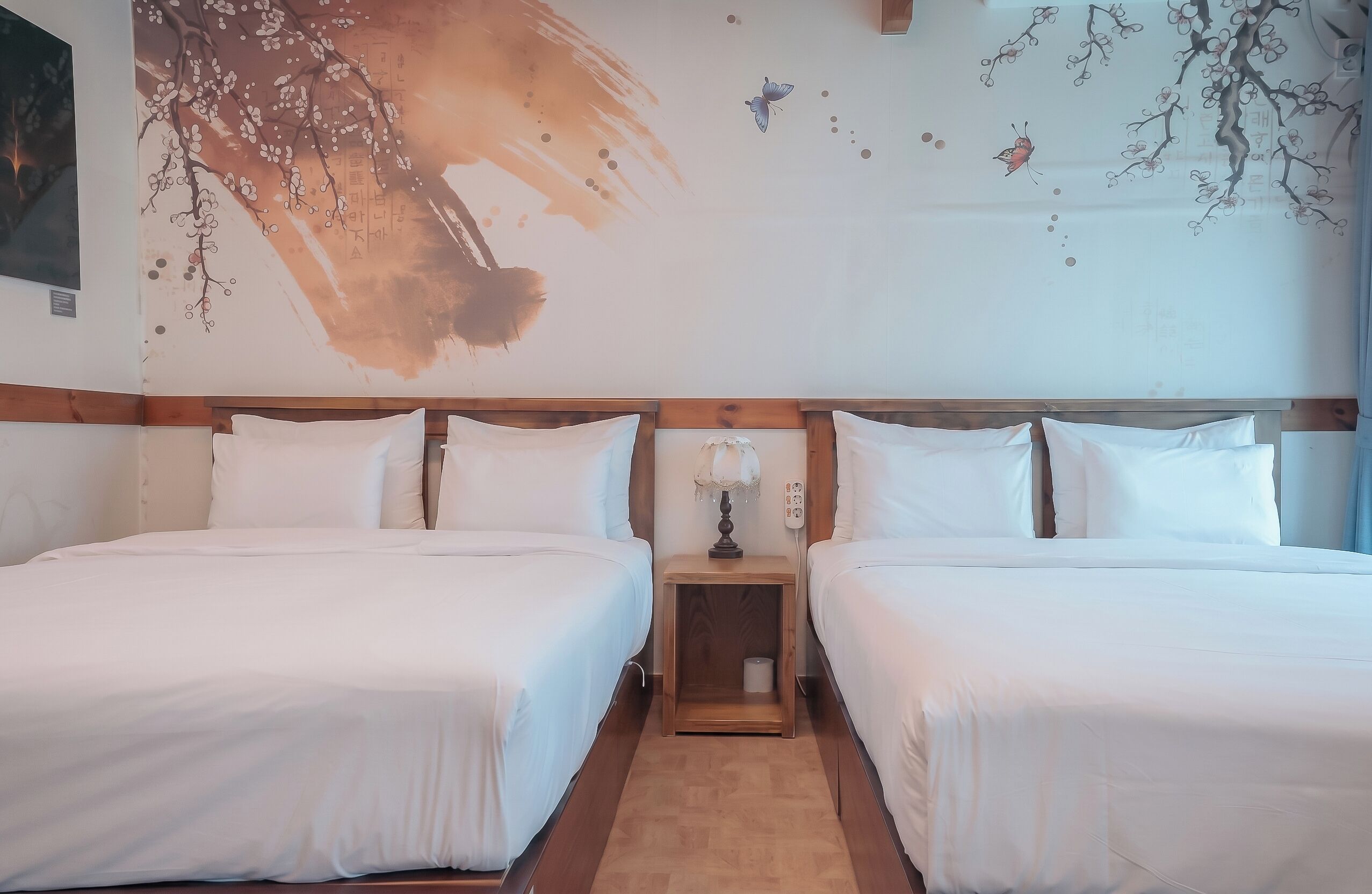 A double room with two beds, wooden headboards, a nightstand with a lamp, and a wall mural with branches and butterflies.