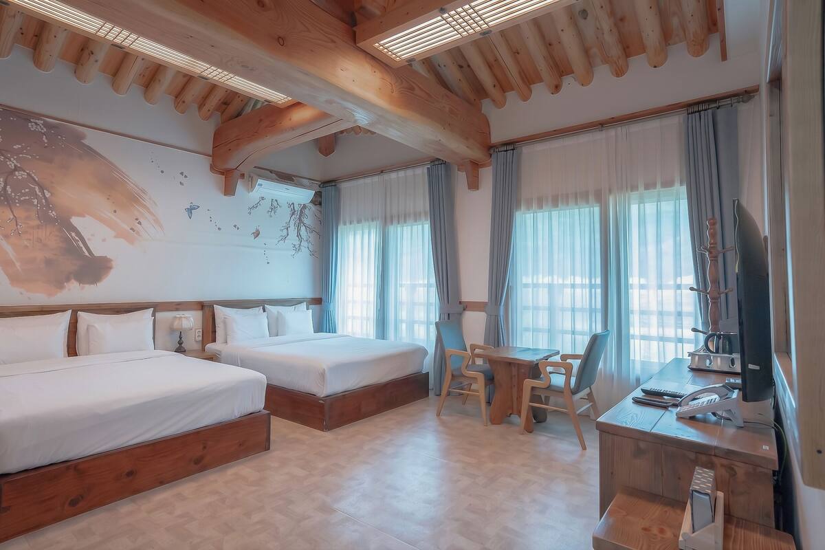 Een hotelkamer met twee bedden, een bureau met stoel, een televisie en grote ramen met gordijnen.