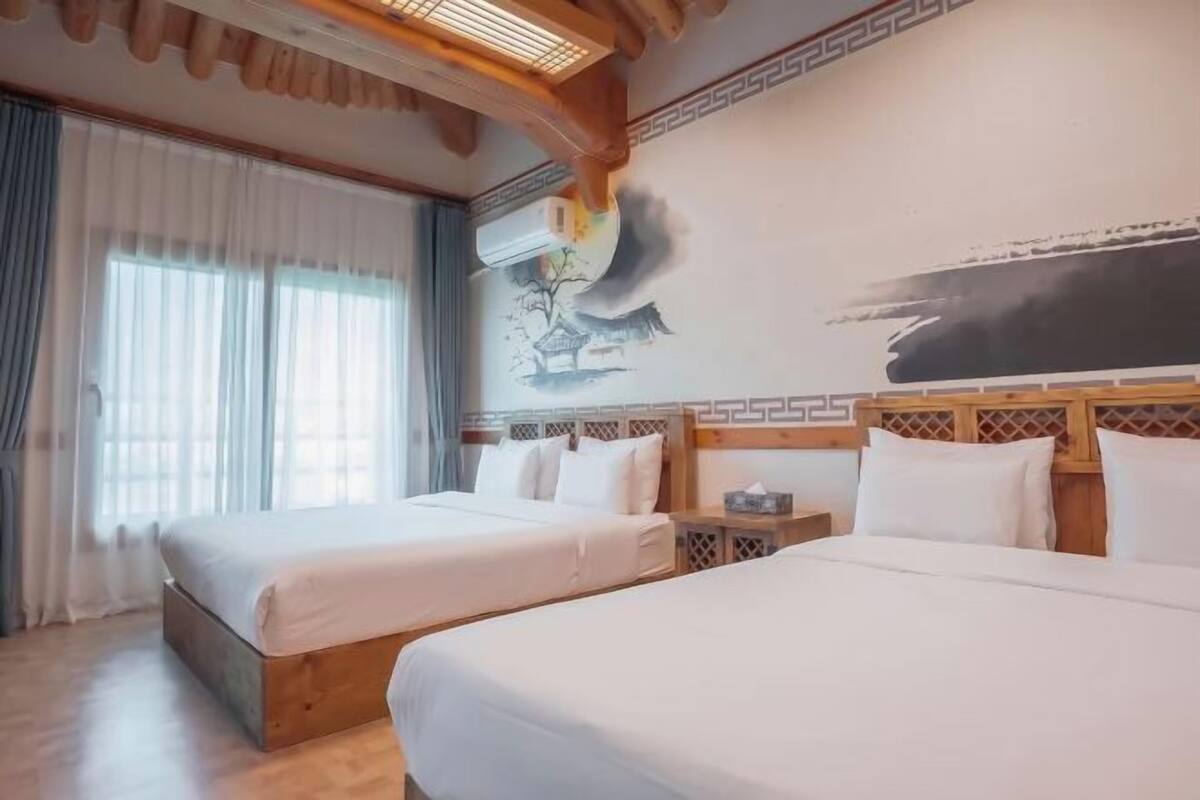 Hotelkamer met twee bedden, een groot raam met gordijnen, een wandairconditioner en een traditionele muurschildering.
