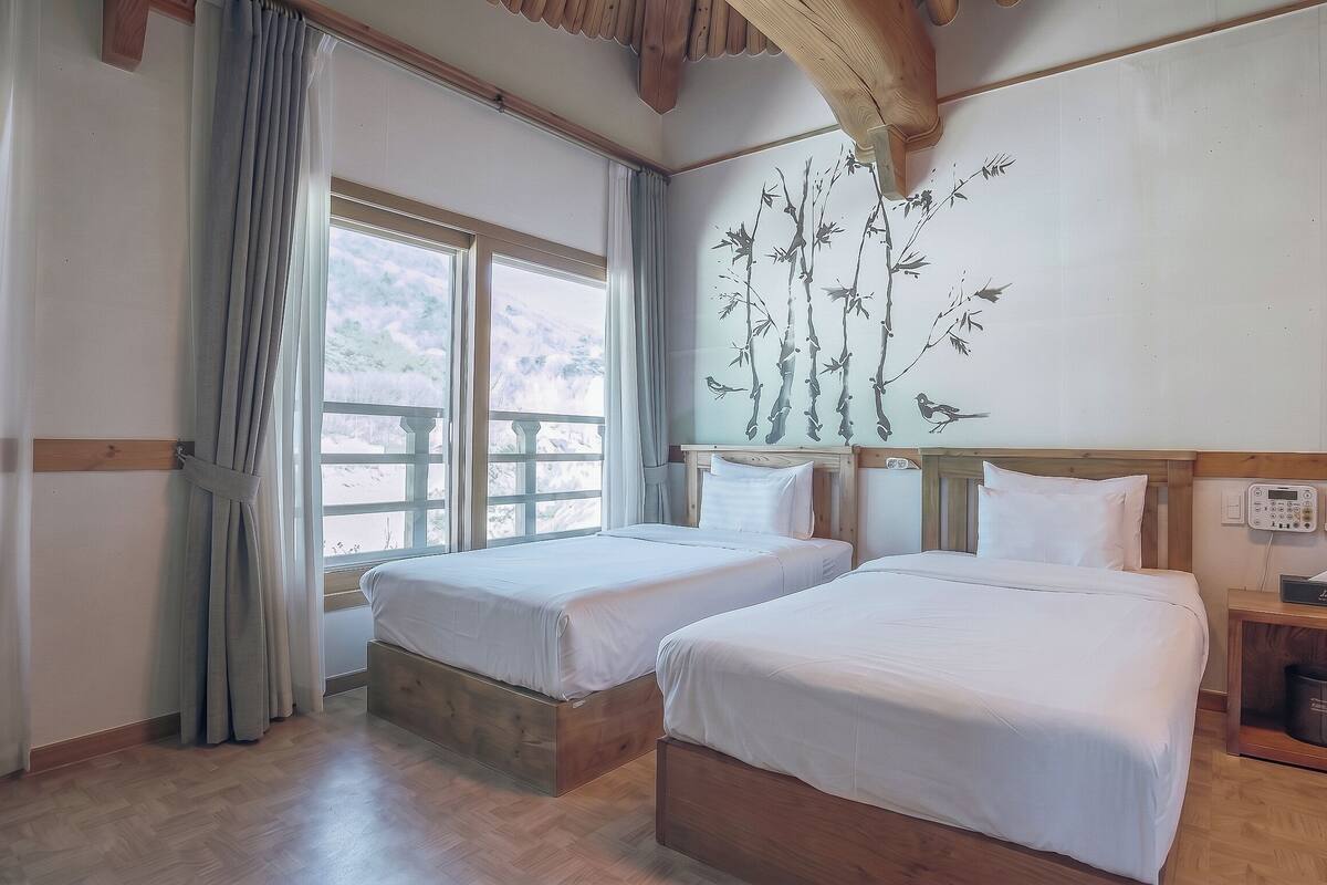 Een hotelkamer met twee bedden, een houten hoofdbord, een raam met gordijnen, een aan de muur bevestigd telefoontoestel en een nachtkastje met zakdoosjes.