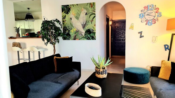 Un salon avec un canapé noir, une table basse et un pouf bleu. On trouve une plante sur la table basse et un tableau au mur.