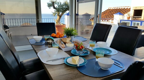 Une table à manger dressée avec des assiettes, des bols et des verres, donnant sur une vue sur la côte, agrémentée d’une plante en pot et d’une terrasse avec des sièges d’extérieur.