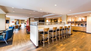 A modern hotel lobby with a central bar area, seating arrangements, and a kitchen visible in the background.