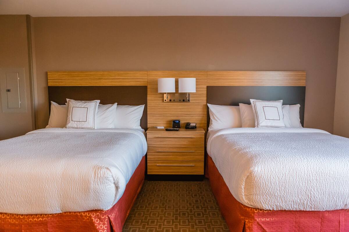 A hotel room with two beds, a wooden headboard, a nightstand with a lamp, and a patterned carpet.