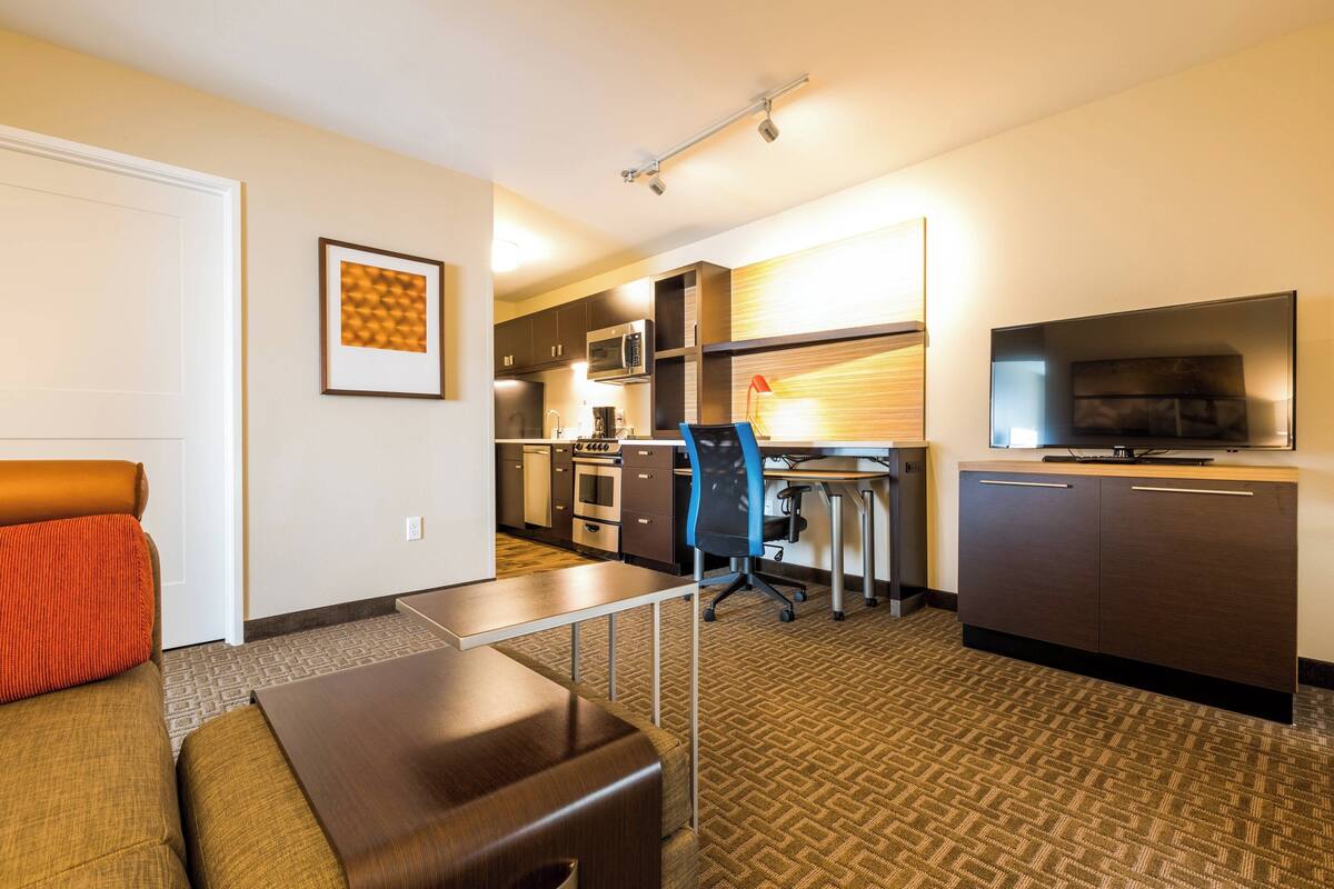 A hotel room with a sofa, a desk, a chair, a television, a microwave, and a kitchen area.