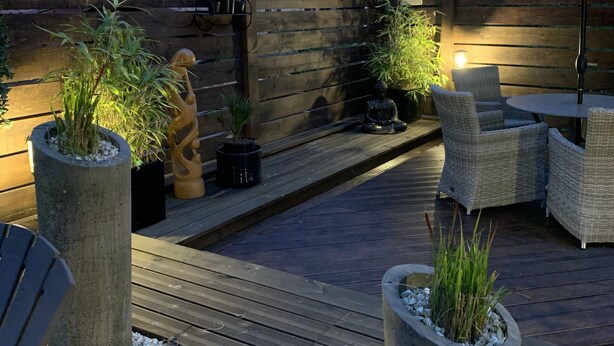 Une terrasse en bois agrémentée de meubles d’extérieur, de plantes en pot et d’une statue de Bouddha.