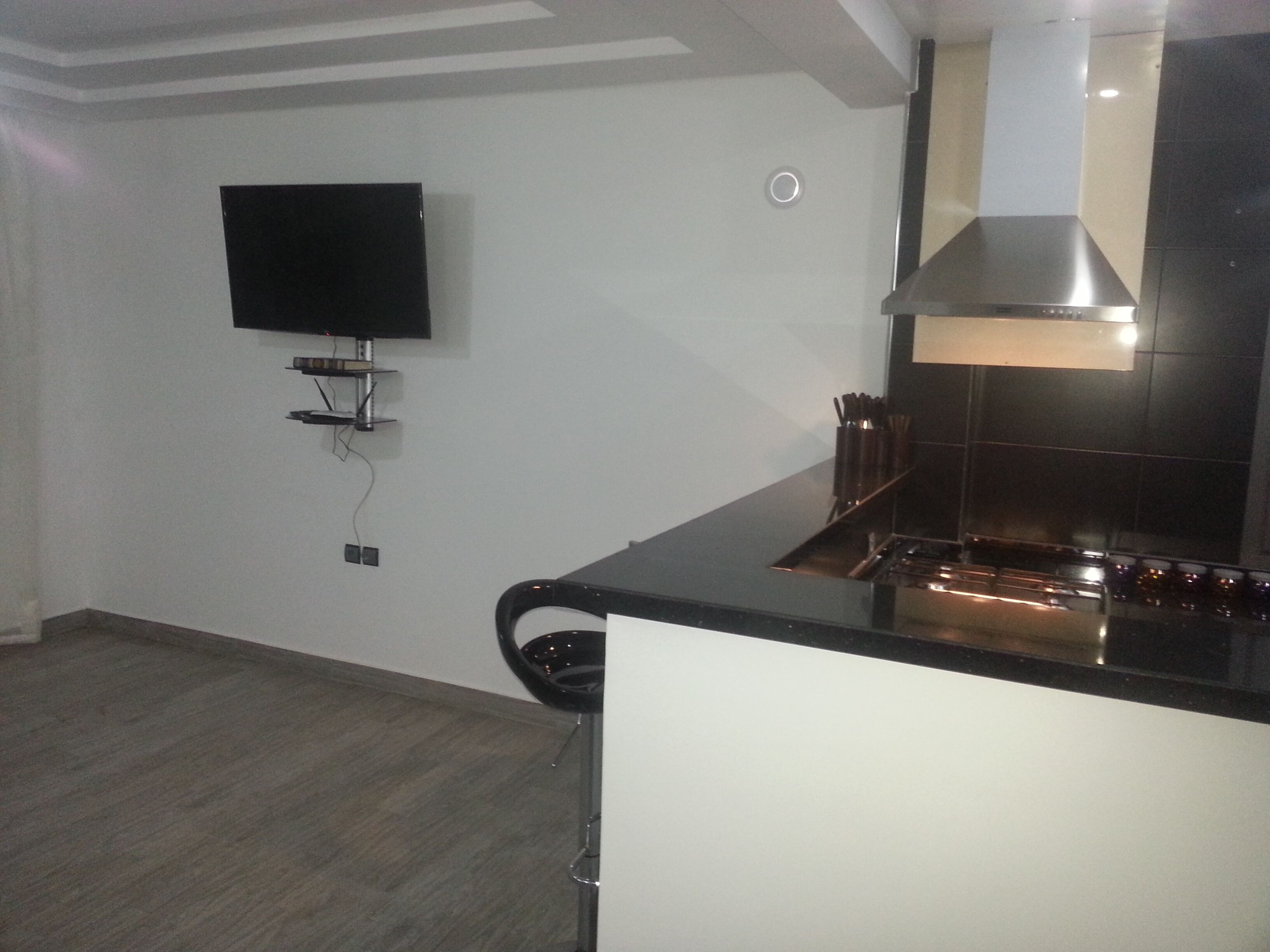 A modern kitchen with a black countertop, stainless steel range hood, and a wall-mounted flat-screen TV.
