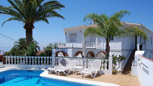 A swimming pool with lounge chairs, a palm tree, and a white building with a tiled roof. - Villa Sandra Top Floor Escape by Casasol (Nerja)