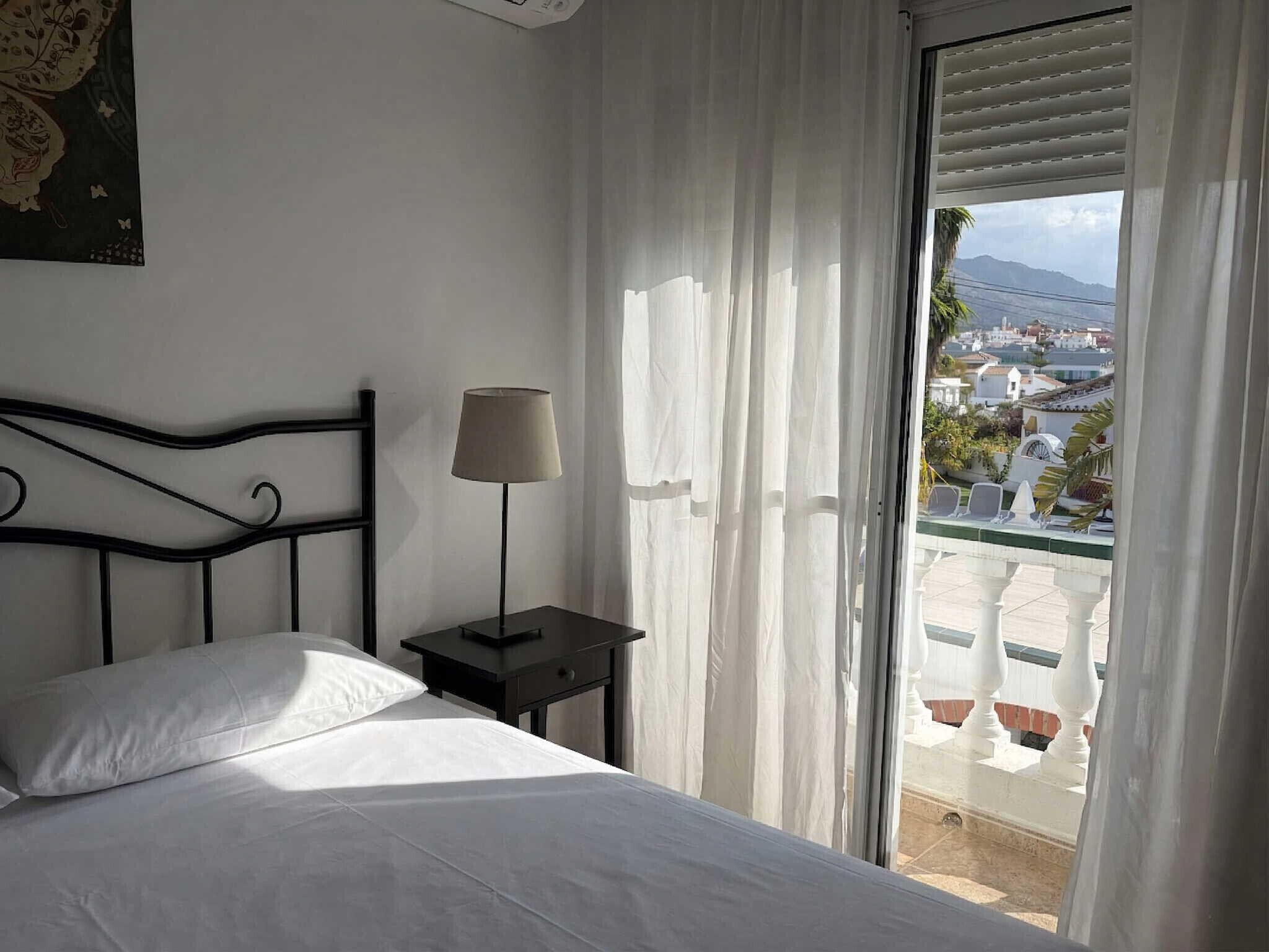 A hotel room with a bed, a nightstand, a lamp, and a view of a balcony with outdoor seating.