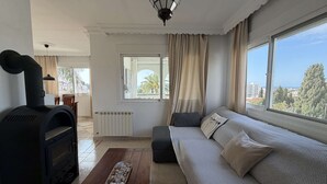 A living room with a fireplace, a sofa, a coffee table with decorative items, a view through a window, and a ceiling light fixture. - Villa Sandra Top Floor Escape by Casasol (Nerja)