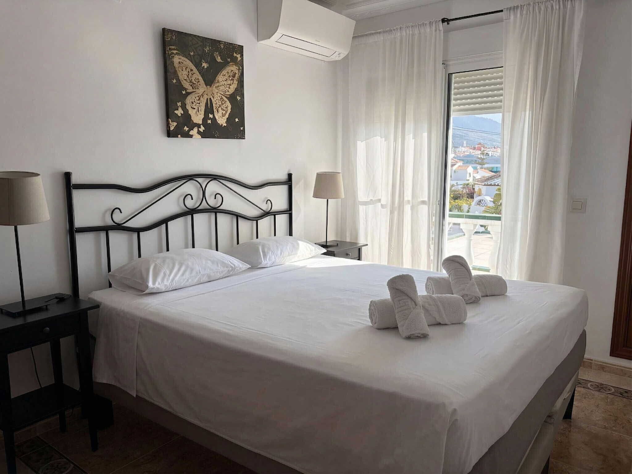 A neatly made bed with white linens, a black metal headboard, two bedside tables with lamps, a window with white curtains, and a butterfly artwork on the wall.