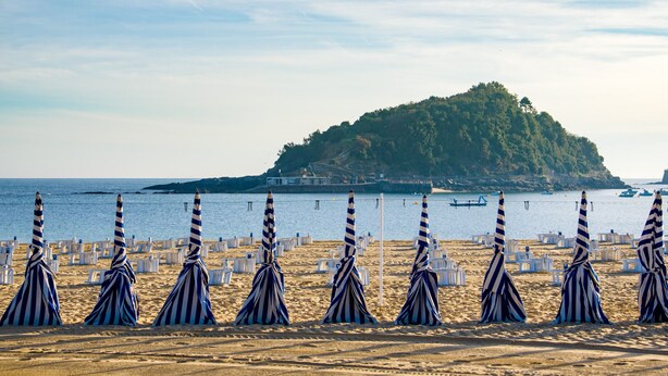 A beach with striped umbrellas and white lounge chairs arranged in rows. - Always Easy I Chillida (San Sebastián/Donostia)