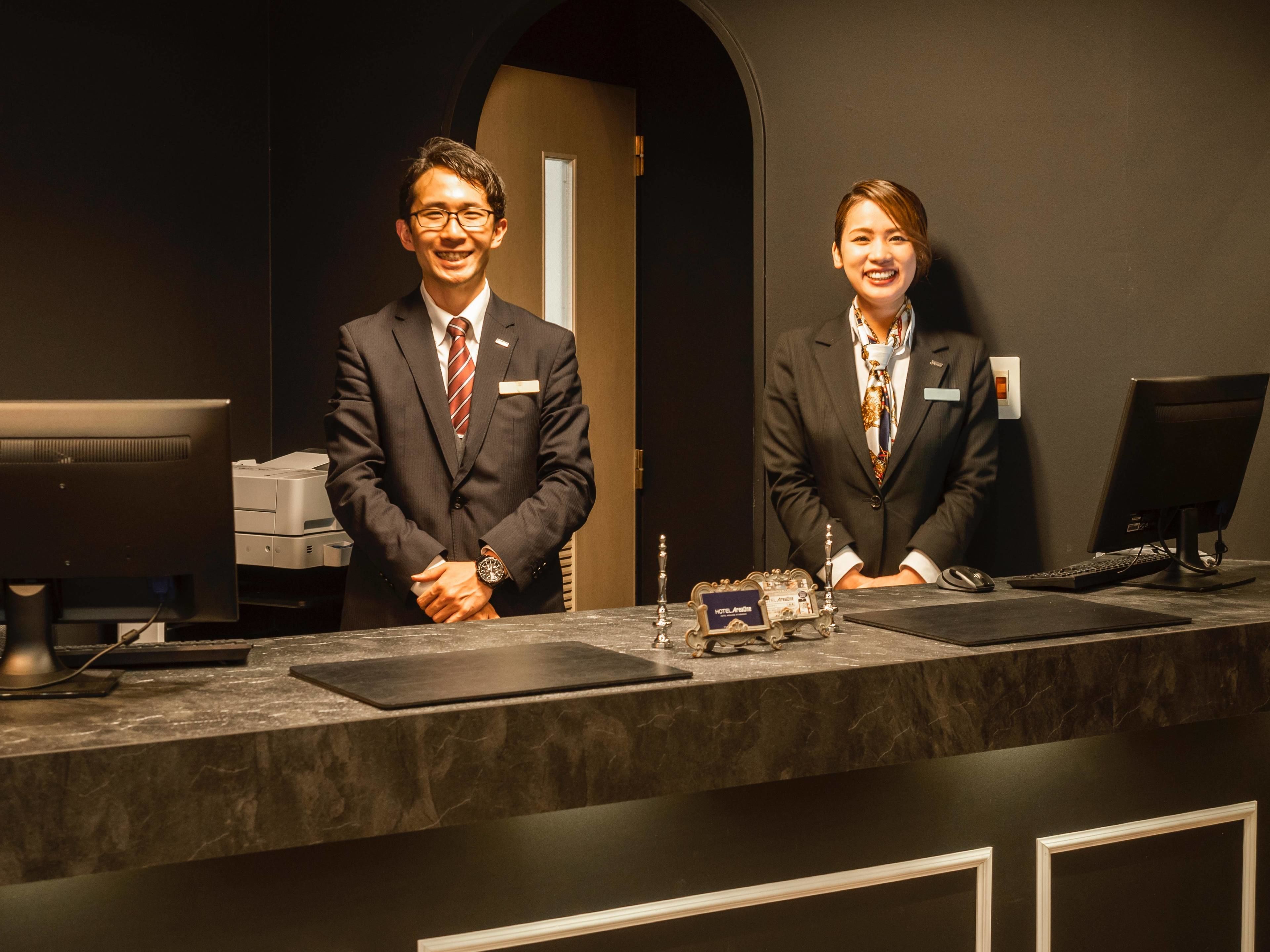 Two individuals at a reception desk with a computer monitor, keyboard, and a small decorative item on the counter.