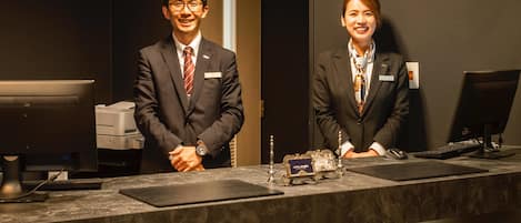 Two individuals at a reception desk with a computer monitor, keyboard, and a small decorative item on the counter.