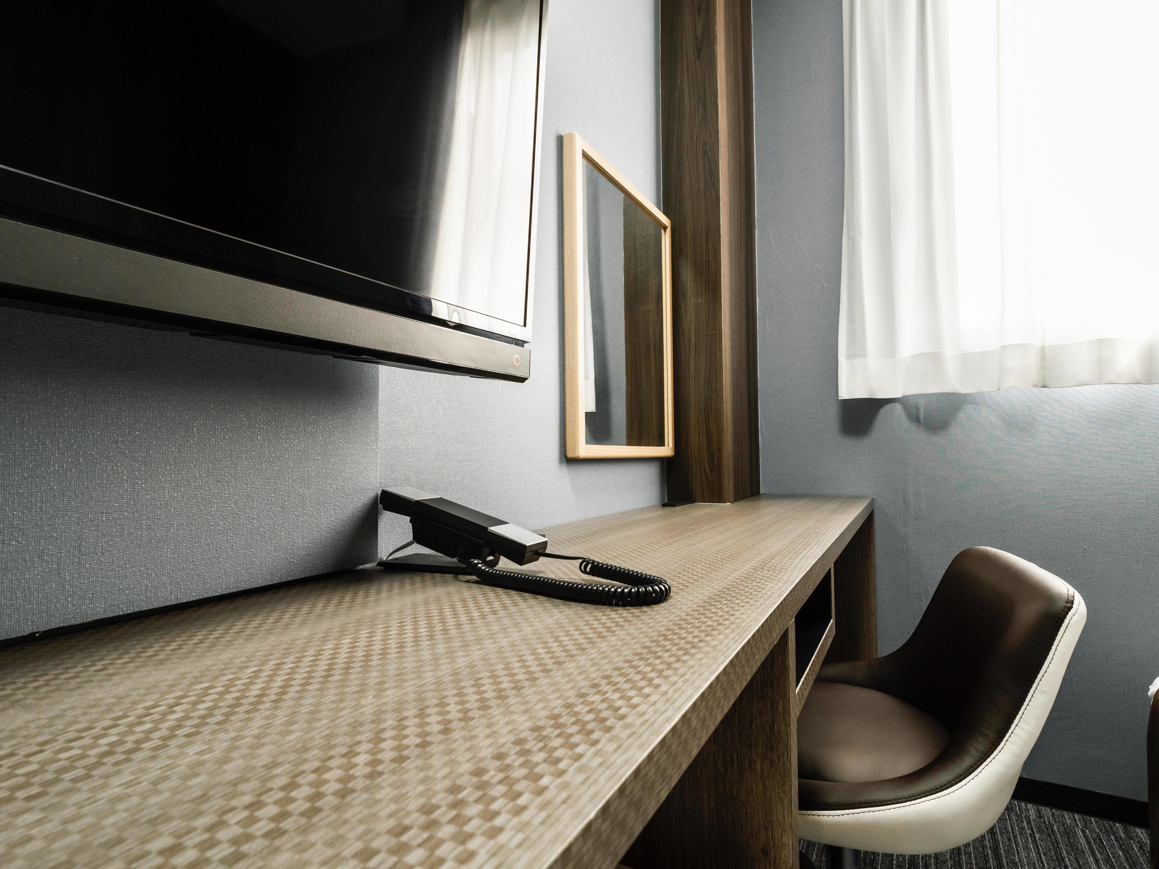 A hotel room with a flat-screen TV mounted on the wall, a desk with a telephone, a chair, and a framed picture on the wall.