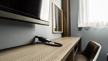 A hotel room with a flat-screen TV mounted on the wall, a desk with a telephone, a chair, and a framed picture on the wall.