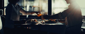 Dos personas brindando con bebidas en una mesa con comida y una jarra de vidrio.