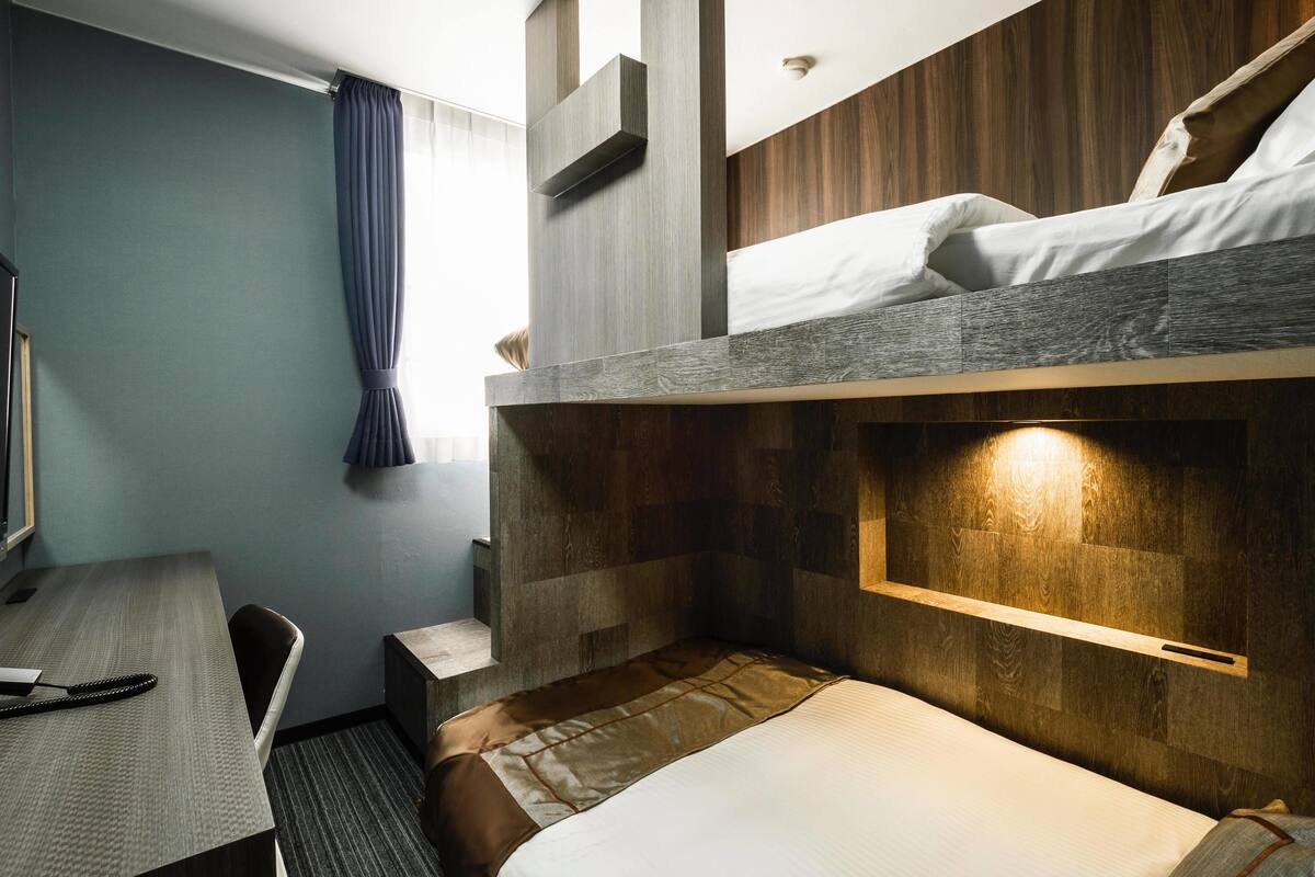 A compact hotel room with a bunk bed, a desk with a chair, and a small seating area.