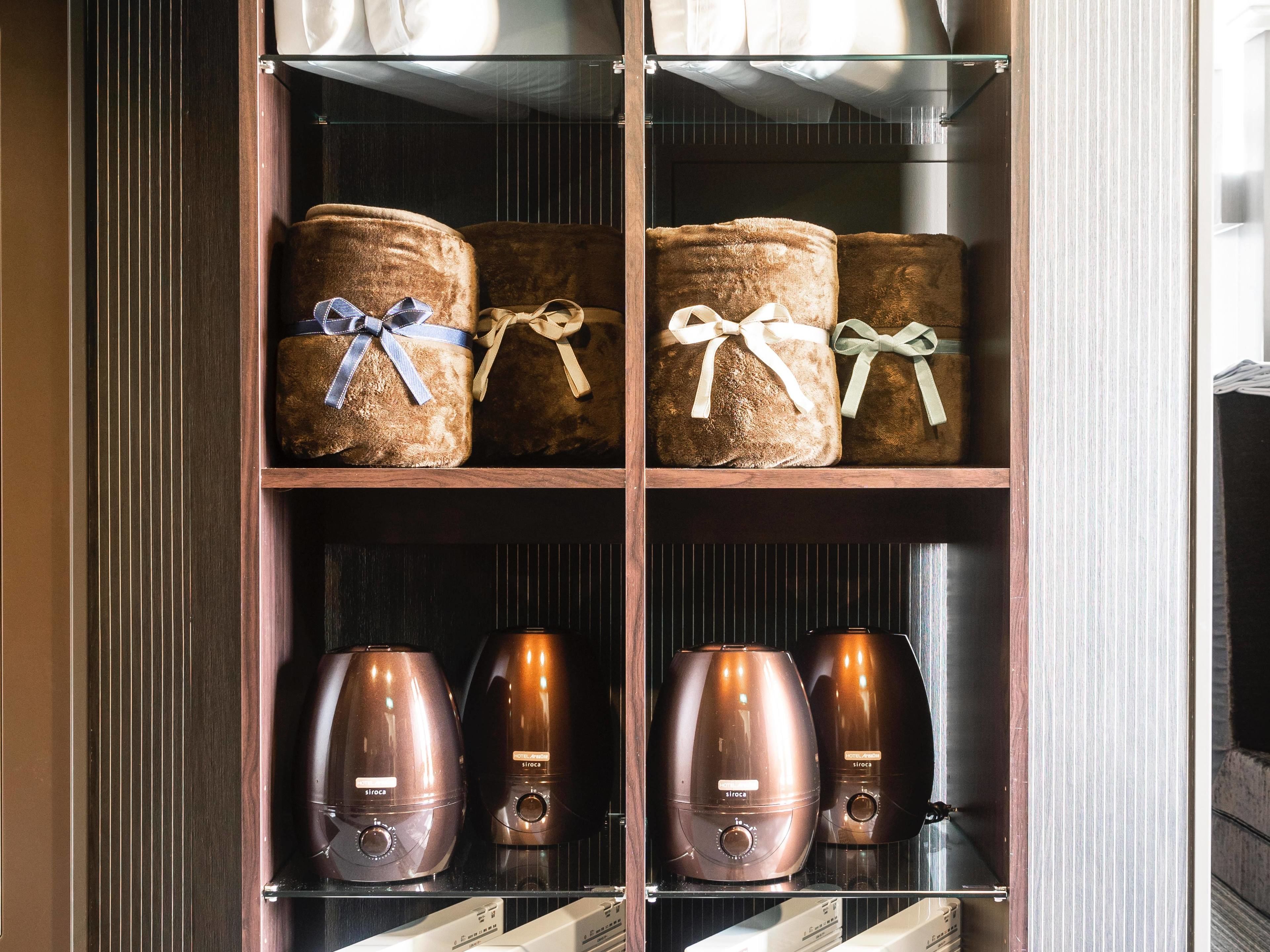 A wooden cabinet with four shelves, each containing items. The top shelf has three burlap sacks tied with ribbons. The middle shelf has two electric kettles. The bottom shelf has two more electric kettles.