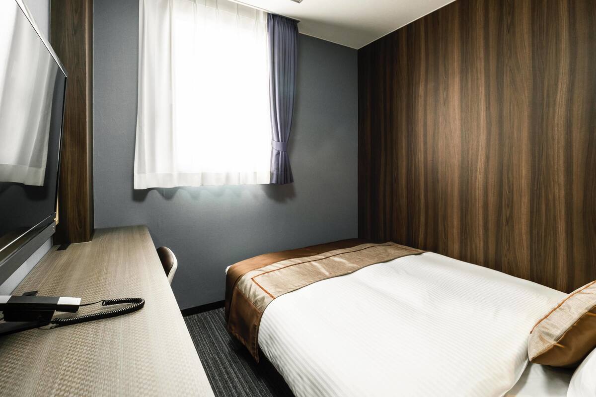 A hotel room with a bed, a desk with a telephone, a window with curtains, and wooden paneling on the walls.