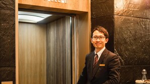 Um homem de terno está em um elevador com painel de madeira e visor digital.