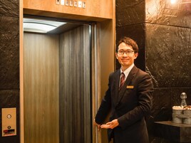 A man in a suit standing in an elevator with a wooden panel and a digital display.