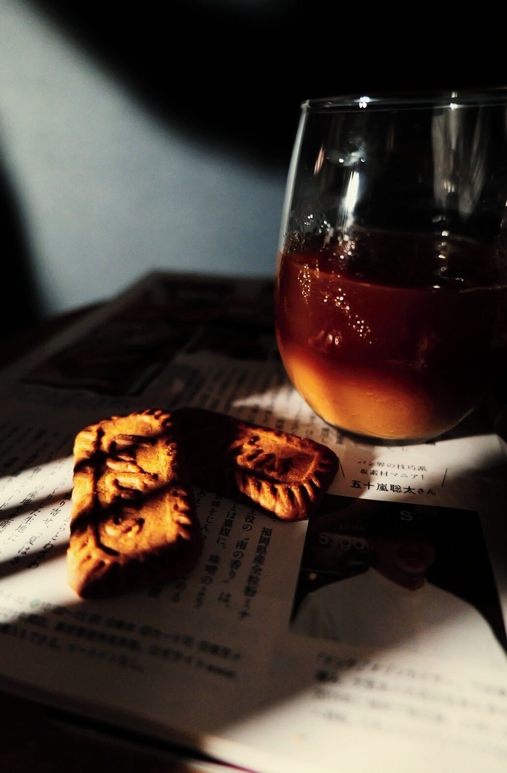 A glass of amber liquid, possibly wine, on a table with a newspaper and two pastries.