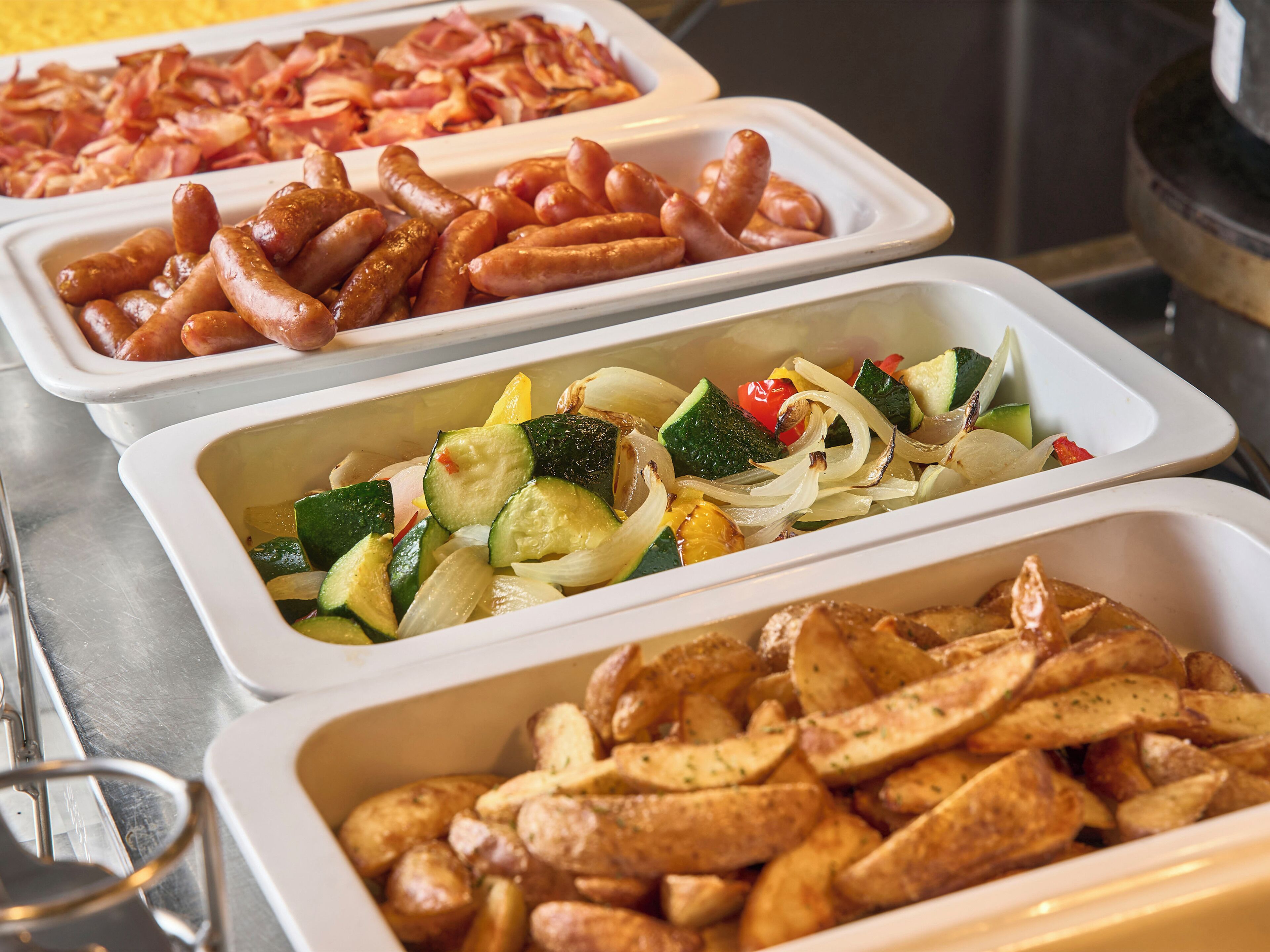 Buffet com diversas opções de comida, incluindo salsichas, legumes e batatas.