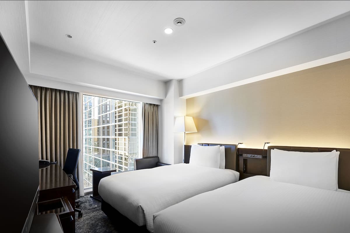 A hotel room with two beds, a desk, a chair, a lamp, and a large window with curtains.