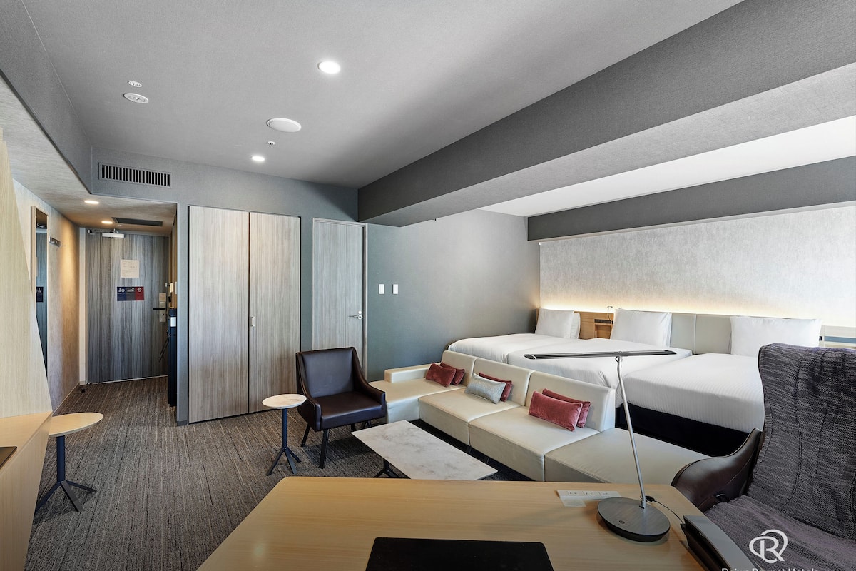 A hotel room with a bed, a desk, a chair, and a sofa. There is a door with a sign, a window, and a ceiling with recessed lighting.