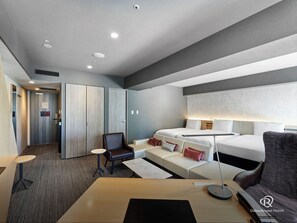 A hotel room with a bed, a desk, a chair, and a sofa. There is a door with a sign, a window, and a ceiling with recessed lighting.