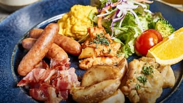 Um prato de comida com linguiça, ovos mexidos, frango grelhado, bacon, batatas assadas, uma salada de alface e tomate, e uma fatia de laranja.