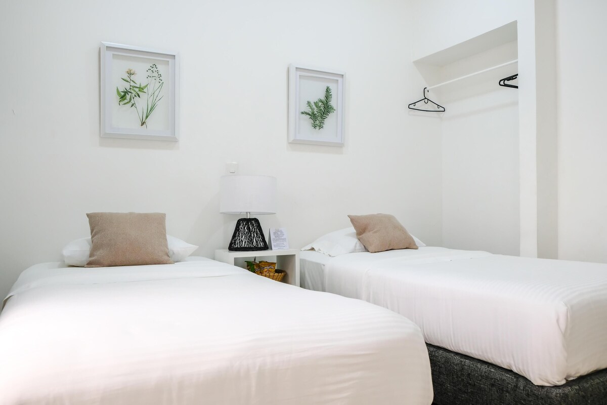 Une chambre d’hôtel avec deux lits, du linge de lit blanc, une table de chevet avec une lampe et deux reproductions botaniques encadrées accrochées au mur.