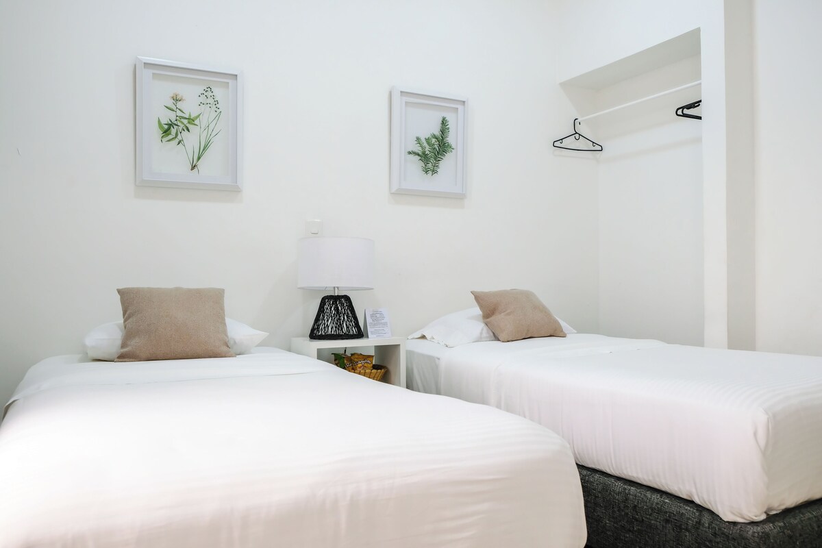 Une chambre d’hôtel avec deux lits, du linge de lit blanc, une table de chevet avec une lampe et deux reproductions botaniques encadrées accrochées au mur.