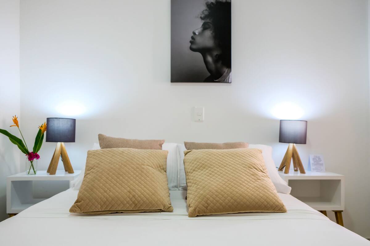 Een keurig opgemaakt bed met twee grote kussens, een nachtkastje met een lamp en een aan de muur hangend schilderij.