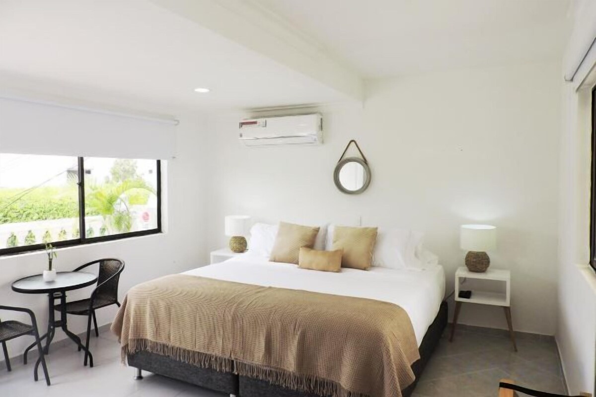Une chambre d’hôtel avec un lit, des tables de chevet, une chaise, une petite table et une fenêtre donnant sur un paysage agréable.