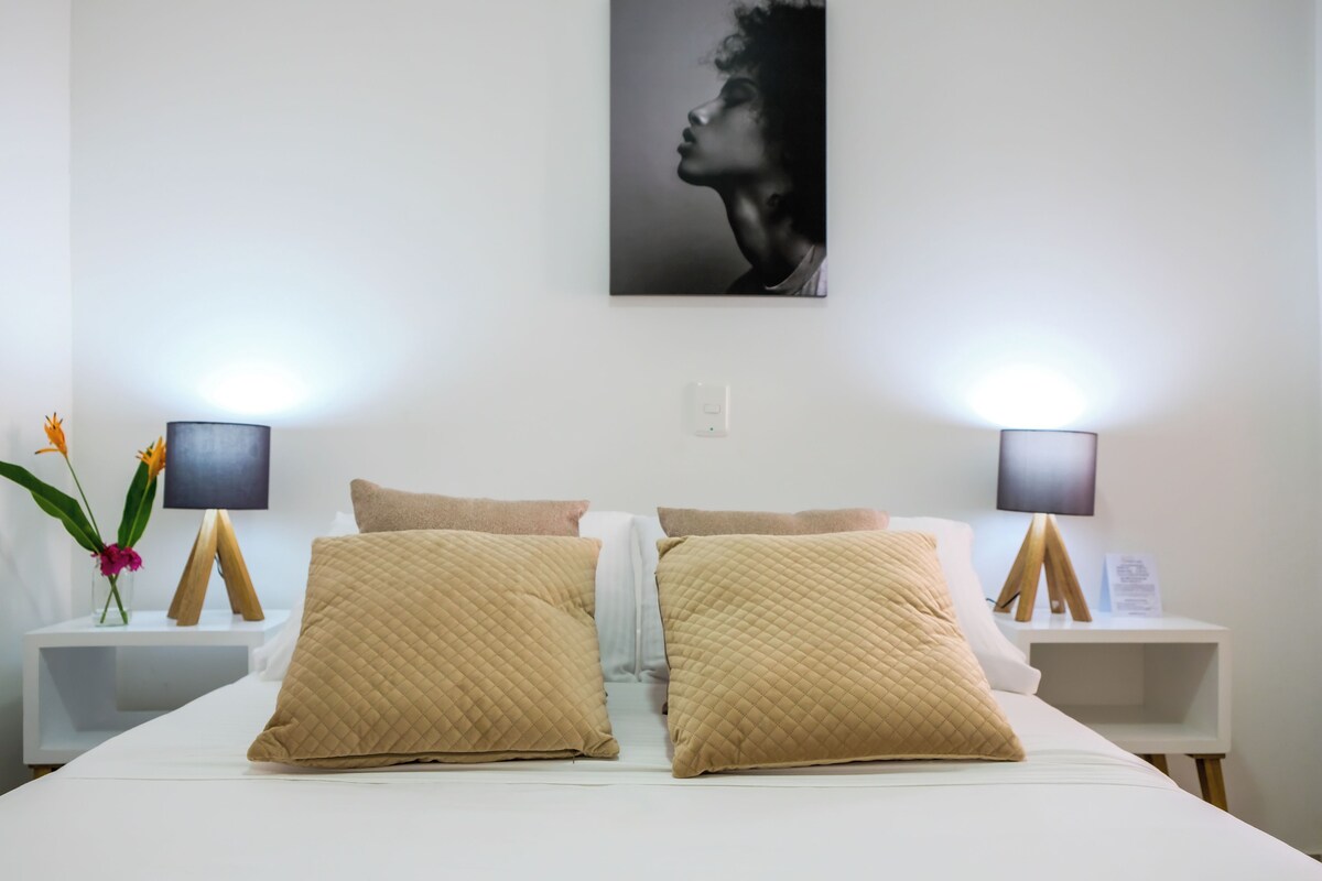 Un lit bien fait, avec deux grands oreillers, une table de chevet avec une lampe et un tableau accroché au mur.